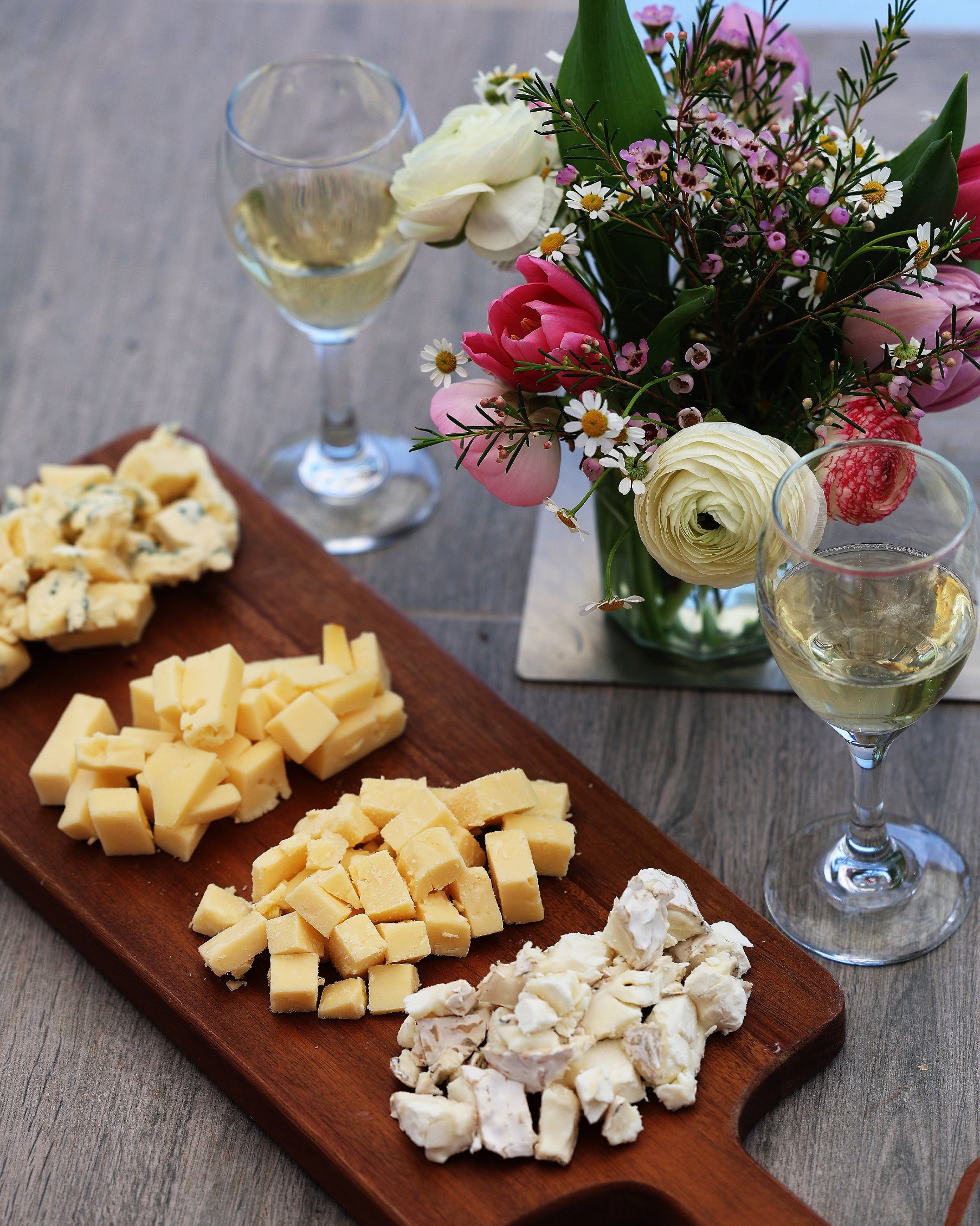 Wooden cheese board with various types of cheese, two glasses of white wine, and a floral arrangement on a wooden table.