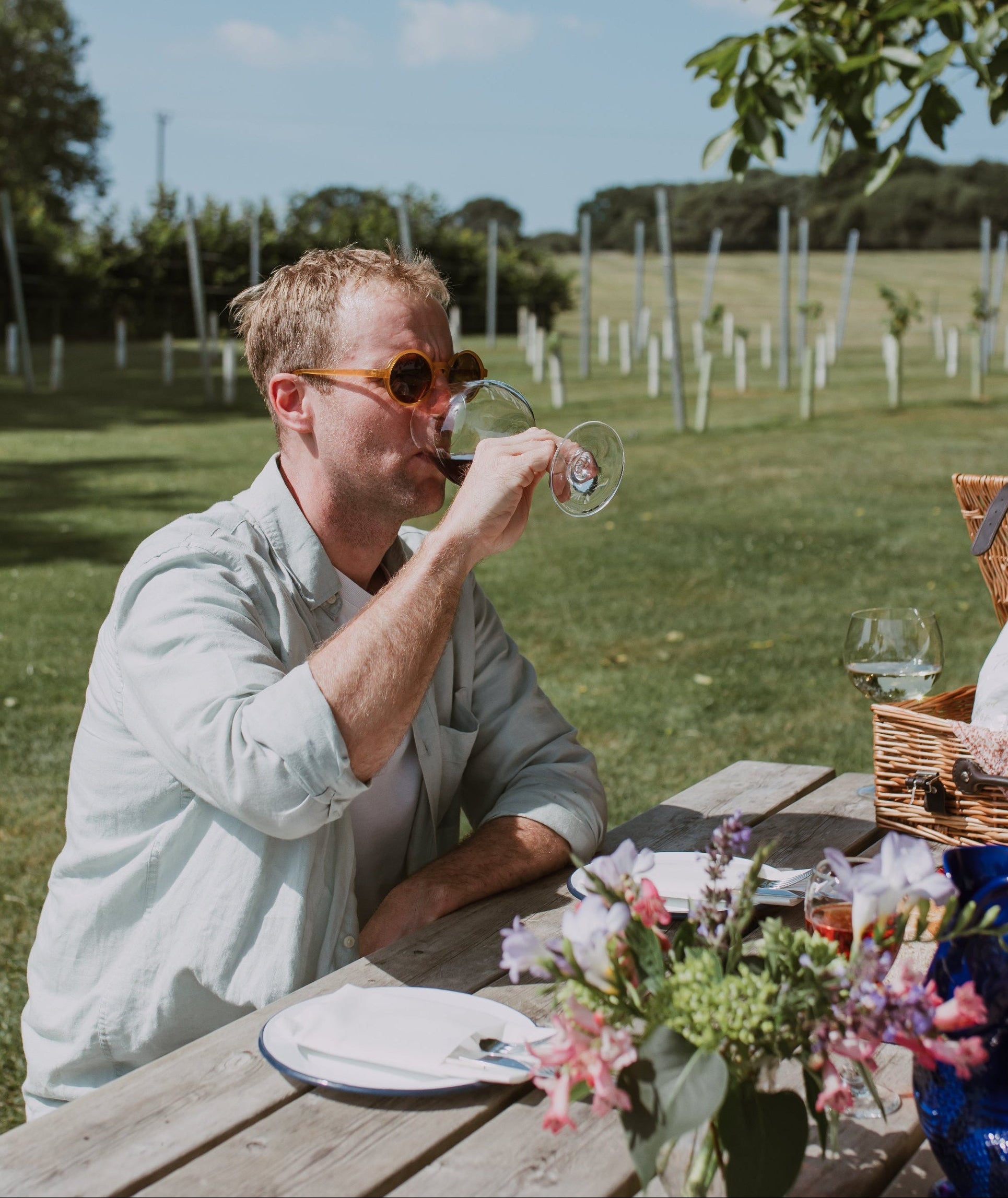 Man drinking from a glass at a picnic table outdoors with a scenic background