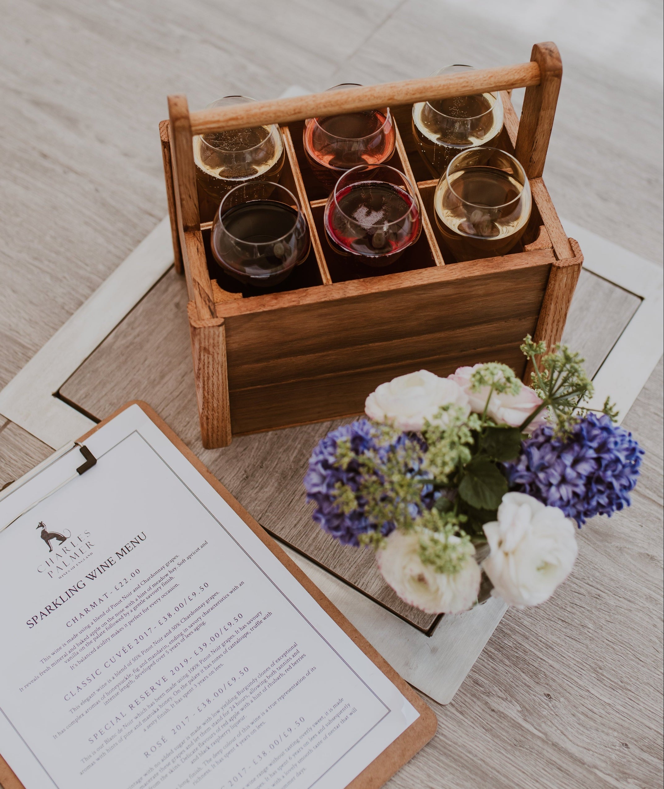 Wooden box with six wine glasses, a floral arrangement, and a menu on a wooden surface.